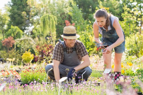 The image depicts two women working together in a well-maintained garden with a lush, green background. They are kneeling on the grass among colorful flower beds that include a variety of blooming flowers in shades of pink, purple, and yellow. One woman, wearing a straw hat and checked shirt, is tending to the flowers with gardening tools, while the other woman, dressed in a blue sleeveless top and denim shorts, is leaning over and appears to be examining or pruning the plants. The garden features a mixture of grass, flower beds, and bordering shrubs, with mature trees in the background providing shade. The natural sunlight highlights the vibrant colors of the flowers and the healthy, dense grass, creating a bright and inviting outdoor scene. This image aligns with gardening and outdoor maintenance services, showcasing the care and attention involved in maintaining a cultivated garden space in the Finsbury Park area, emphasizing practical, hands-on gardening work.