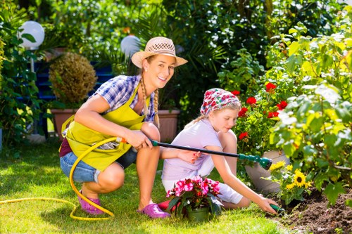A woman and a young girl are gardening together in a well-maintained backyard garden with lush green foliage and colorful flowers. The woman, dressed in a checked shirt, pink wellington boots, and a wide-brimmed straw hat, is crouched beside the girl, holding a yellow garden hose, and smiling as she observes the child watering a flower bed. The girl wears a floral patterned sun hat, a light purple t-shirt, and pink shoes, and is focused on carefully watering the flowering plants, which include bright red, yellow, and pink blooms. The garden features a neatly cut lawn, landscaped flower beds with rich soil, and a wooden border separating the grass from the flower areas. In the background, there are decorative white lanterns hanging from tall trees and dense shrubbery, creating a vibrant outdoor setting under natural daylight. The scene captures a moment of outdoor gardening, emphasizing the nurturing of plants and engagement with nature, reflecting professional gardening and landscaping practices that Lawn Mowing Finsbury Park might provide in this South London area postcode.