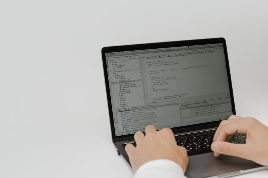 A person's hands are positioned on a laptop keyboard, with the screen displaying a code editor or development environment. The laptop is placed on a plain, light-colored surface, and the background is