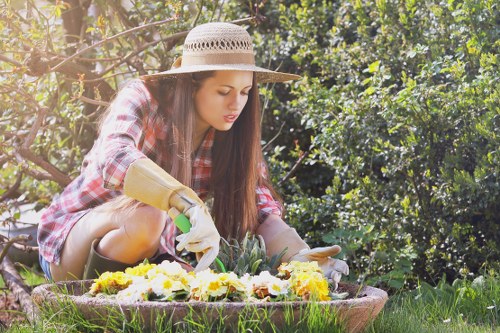 A young woman in a garden during daytime, wearing a wide-brimmed straw hat, a red checked shirt, and gardening gloves, is tending to flower beds with a focused expression. She is kneeling on a lush, well-maintained grassy lawn with vibrant flowering plants arranged in a circular bed, bordered by soil and small stones. Behind her, dense green bushes and trees provide a natural backdrop, with sunlight filtering through the foliage, creating a warm and peaceful outdoor environment. The scene illustrates typical gardening activities in a landscaped outdoor space, possibly associated with local garden maintenance or landscaping services in Finsbury Park, contributing to the overall aesthetic and health of the outdoor area.